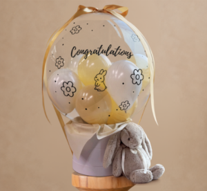 A hot air balloon style gift box featuring a clear balloon filled with smaller white and cream balloons, printed with "Congratulations", flowers, and a small bunny illustration, paired with a plush plush bunny sitting next to the box.