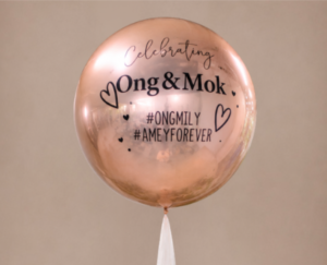 A spherical metallic rose gold balloon customized with black text reading "Celebrating Ong & Mok" along with hand-drawn hearts and event hashtags.