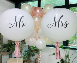 Two large white balloons printed with cursive script reading "Mr" and "Mrs", decorated with pink tassels and surrounded by a cluster of rose gold and white balloons.