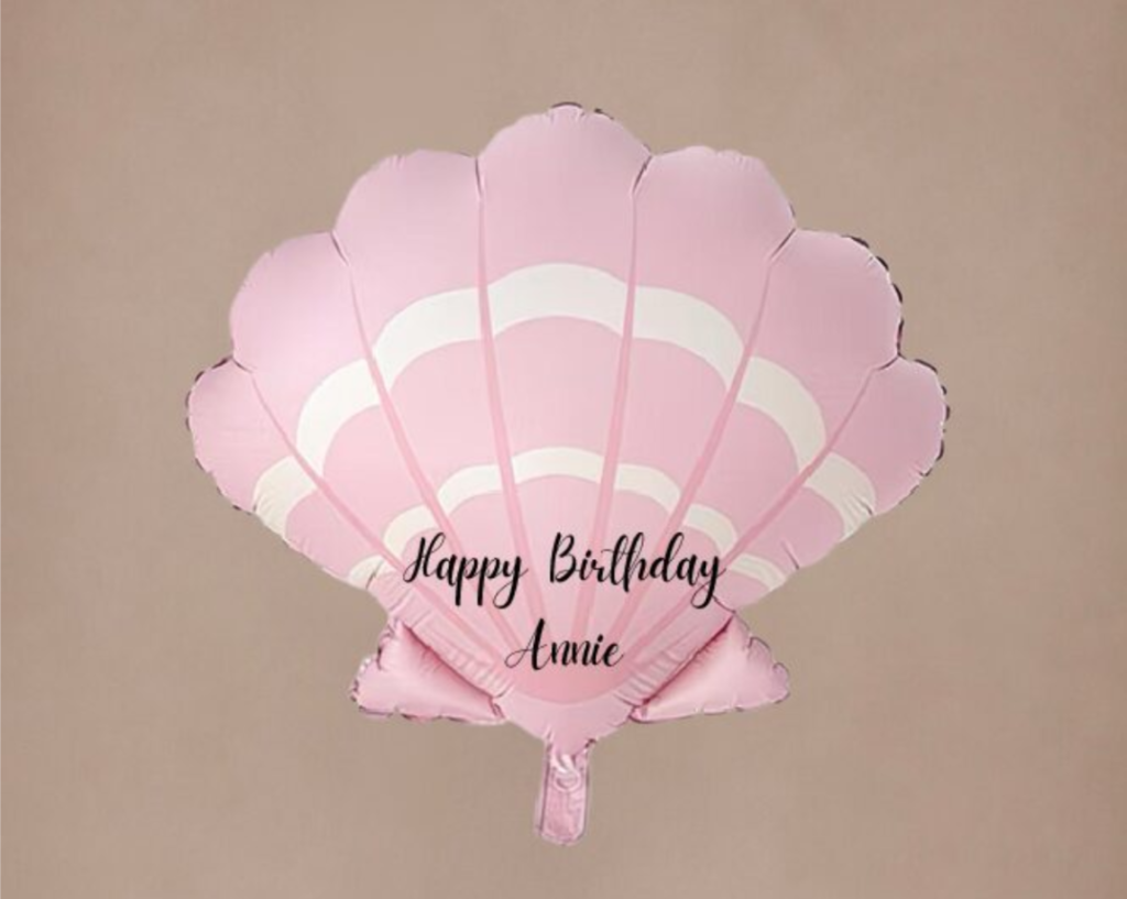 A seashell-shaped pink foil balloon printed with black cursive text reading "Happy Birthday Annie".